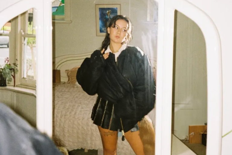 Australian musician Mallrat reflected in a vanity mirror wearing a dark jacket in a promotional photo captured by Meg Siejka.